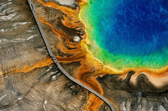 SOURCE CHAUDE DU GRAND PRISMATIC, PARC NATIONAL DE YELLOWSTONE, WYOMING, ÉTATS-UNIS (44° 31’ N – 110° 50’ O). Situé sur un plateau volcanique qui chevauche les États du Montana, de l’Idaho et du Wyoming et créé en 1872, Yellowstone est le premier et le plus ancien parc national du monde. Alors que les États-Unis achevaient la « conquête de l’Ouest » et massacraient les derniers bisons, certains eurent l’intuition que la nature devait être protégée. Le parc s’étend sur 9 000 km2 et présente la plus grande concentration de phénomènes géothermiques du globe, avec plus de 300 geysers, et des milliers de fumerolles et sources chaudes. D’un diamètre de 112 m, le Grand Prismatic est le bassin thermal le plus vaste du parc, et le troisième au monde par sa taille. Le spectre de couleurs qui lui a valu son nom est dû en son centre à la profondeur de l’eau et, à la périphérie de la vasque, à la présence d’algues microscopiques dont la croissance dans l’eau chaude, diffère en fonction de la température. Réserve de la biosphère depuis 1976, inscrit sur la liste du patrimoine mondial de l’Unesco en 1978, le parc national de Yellowstone reçoit près de 4 millions de visiteurs chaque année, essentiellement entre les mois de mai et d’octobre. Le continent nord-américain, où sont situés les cinq sites naturels les plus fréquentés du monde, a accueilli plus de 137 millions de touristes internationaux en 2017, soit plus de 10 % des 1,326 milliard de touristes internationaux comptabilisés cette année-là. À l’échelle du monde, l’activité touristique représente plus de 10 % du PNB mondial. S’il représente 277 millions d’emplois directs, indirects et induits, l’industrie du tourisme et des voyages impacte négativement l’environnement, les ressources naturelles et les cultures locales.